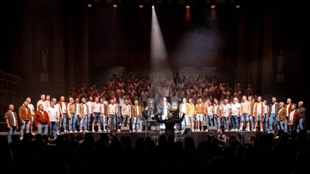 The London Gay Men's Chorus standing on a vast stage with a spotlight performing in front of an audience.