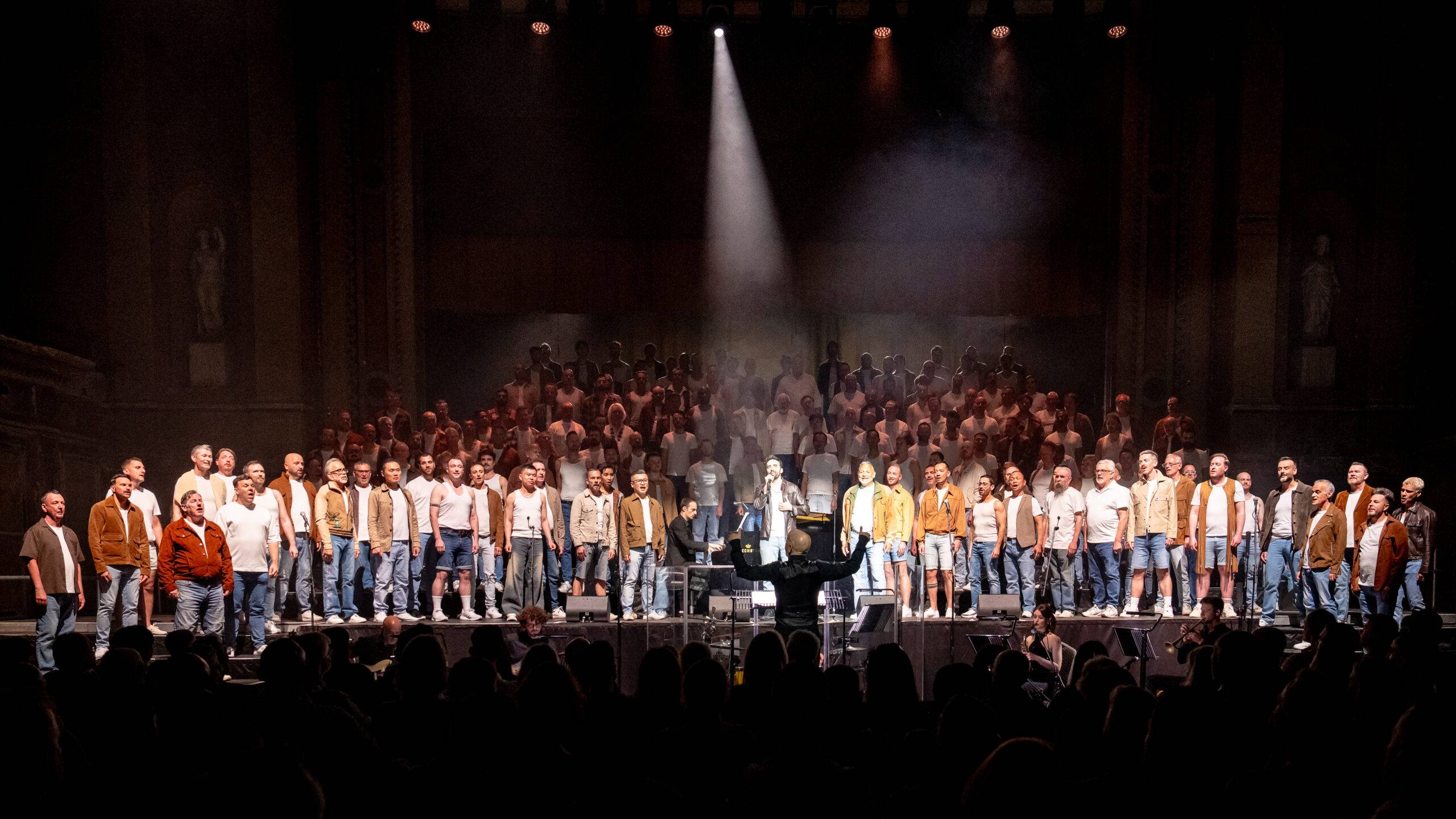 The London Gay Men's Chorus standing on a vast stage with a spotlight performing in front of an audience.